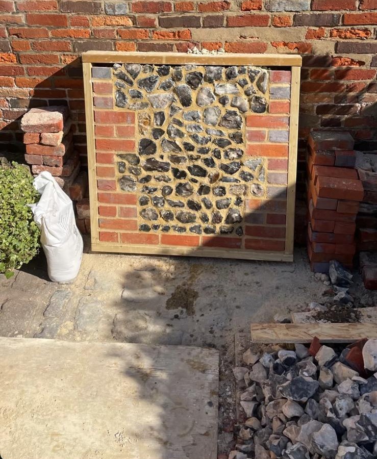 Framed sample panel of dark knapped flint in yellow mortar with red brick edging, propped against an old brick wall beside stacked bricks and loose flints