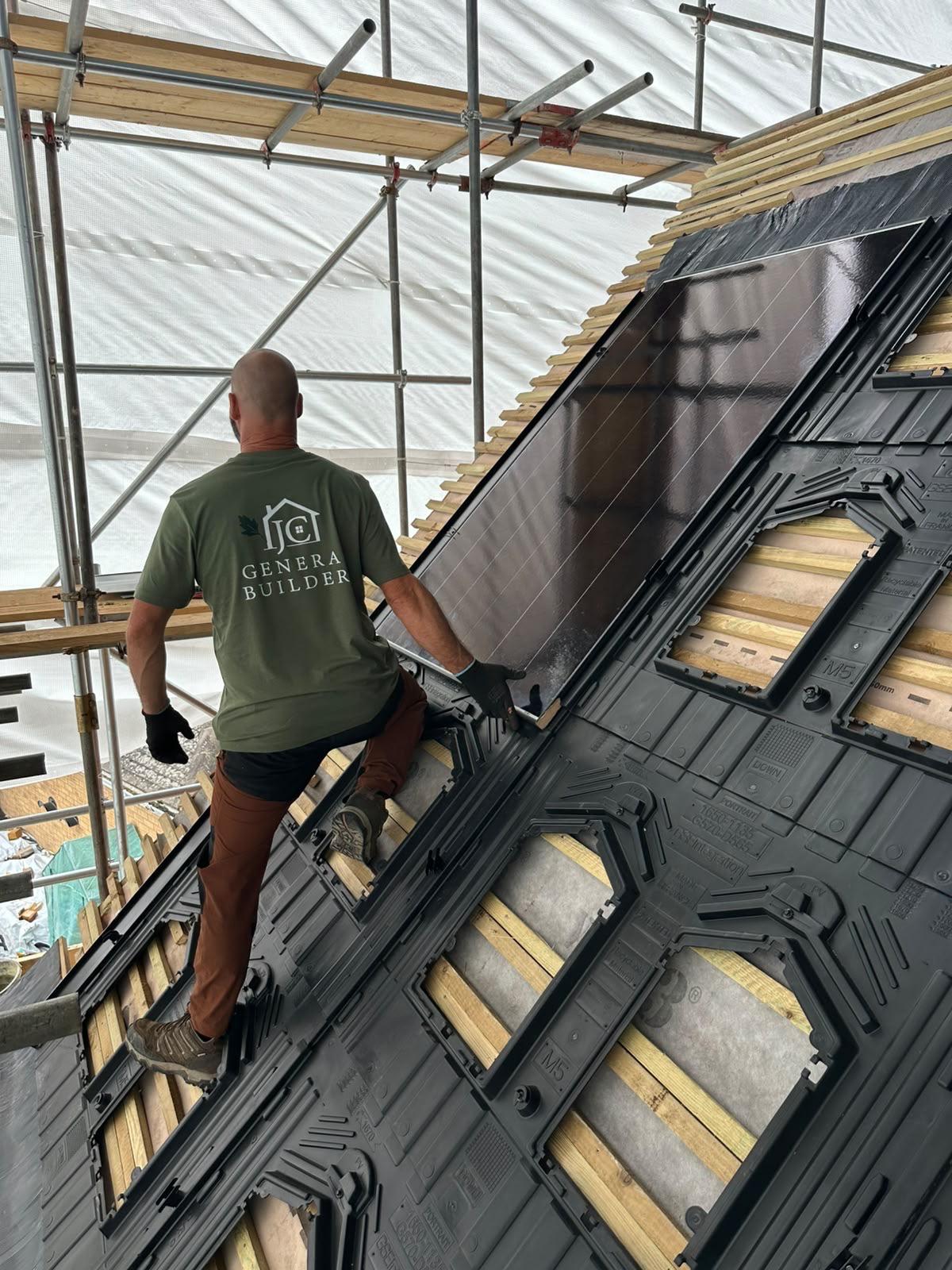 Builder in a green J.C General Builders t-shirt lowering a black solar panel into a plastic in-roof mounting tray on a battened roof under scaffolding