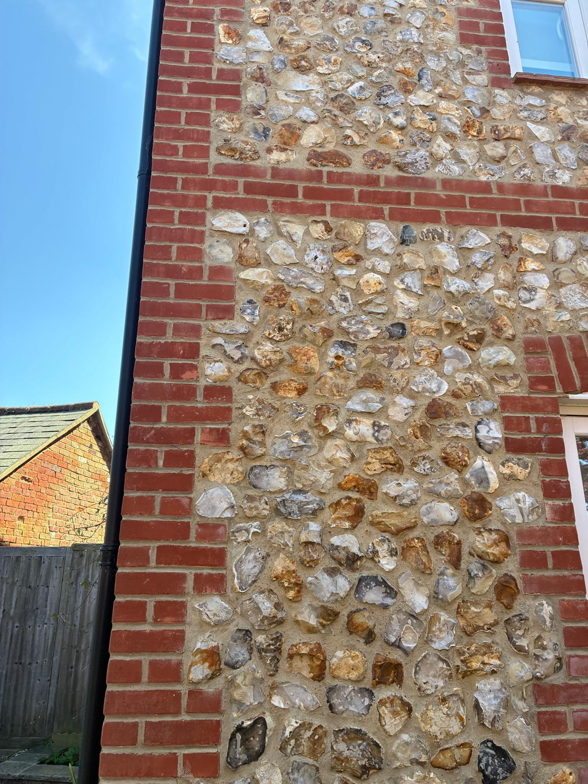 Two-storey wall of knapped flint set in lime-coloured mortar, framed by red brick quoins and a horizontal brick band, with a black downpipe at the corner