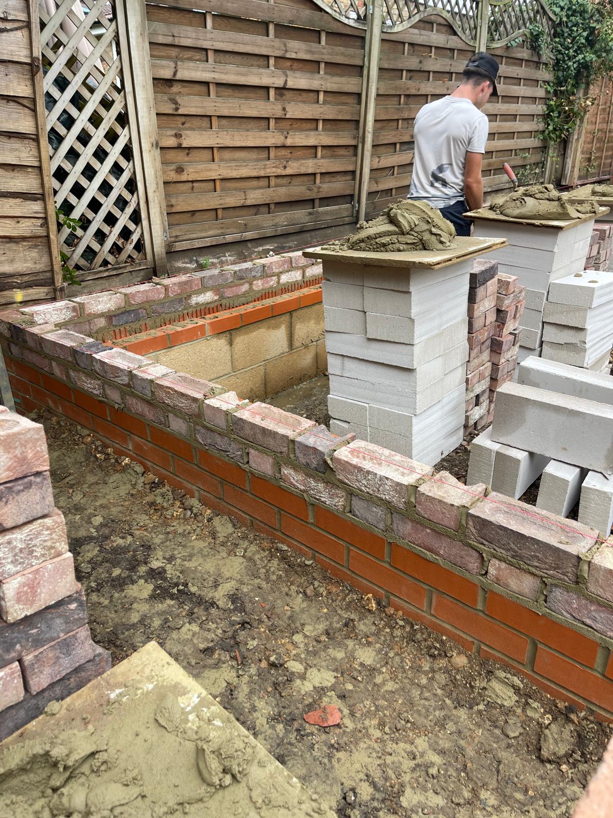 Builder laying reclaimed bricks on a raised planter with a red brick lower course, blockwork behind and mortar boards on block stacks