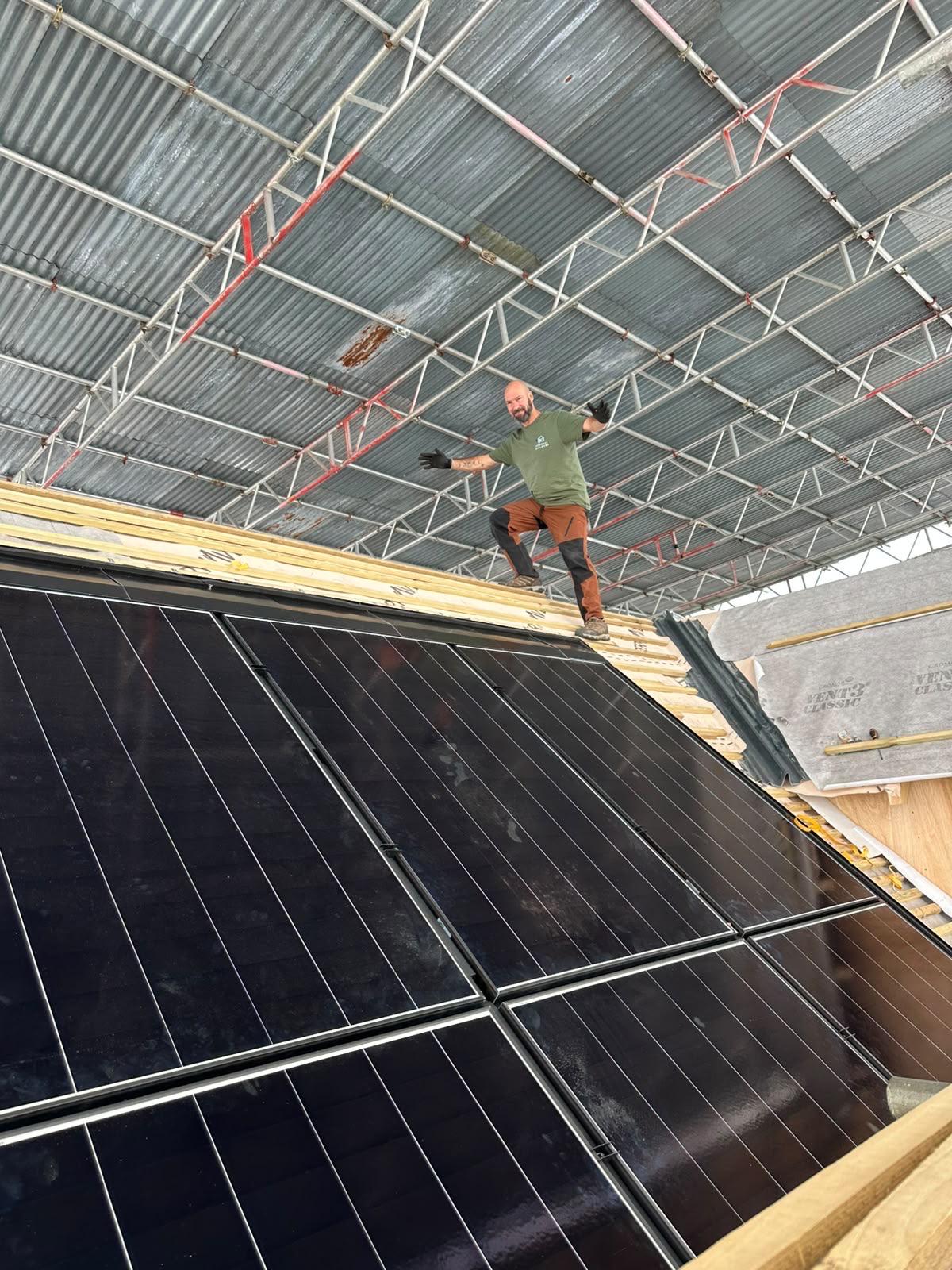 Builder in a J.C General Builders t-shirt standing on roof battens above a row of black in-roof solar panels, under a scaffold canopy