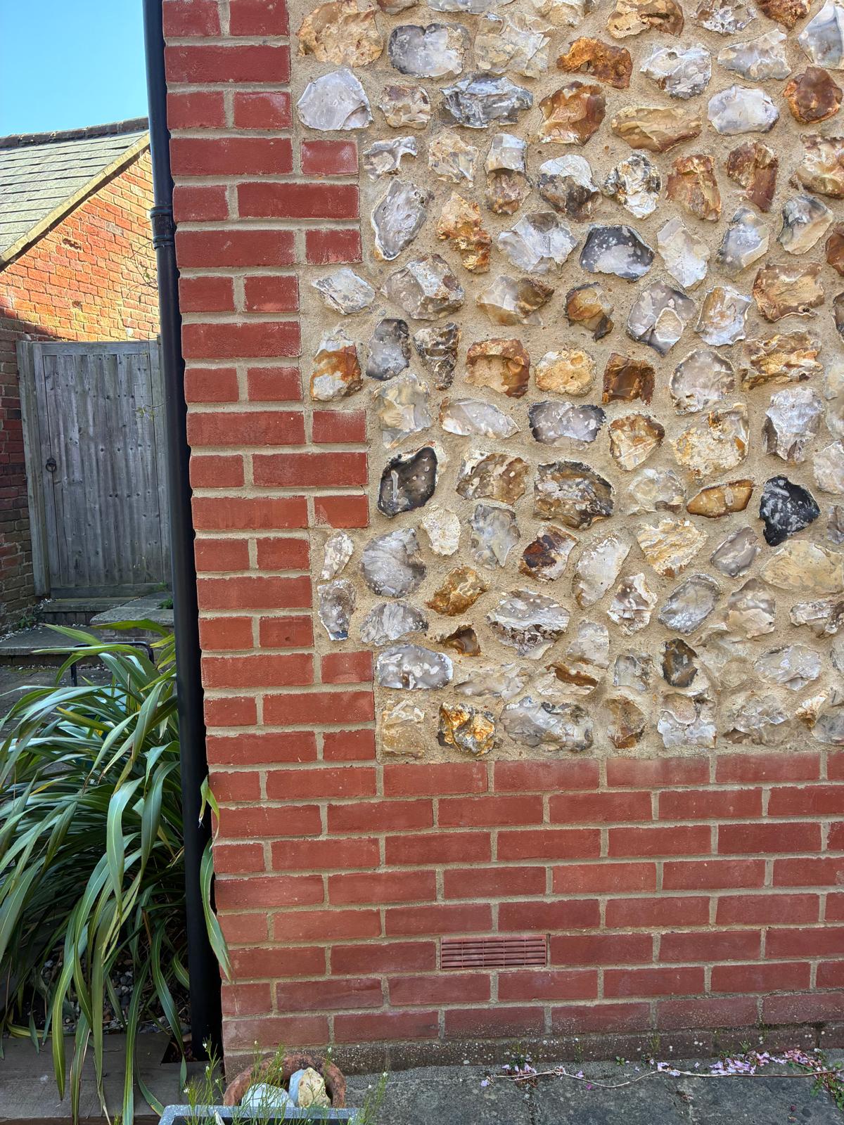 Close-up of a knapped flint panel above a red brick plinth with an air brick, edged by toothed red brick quoins